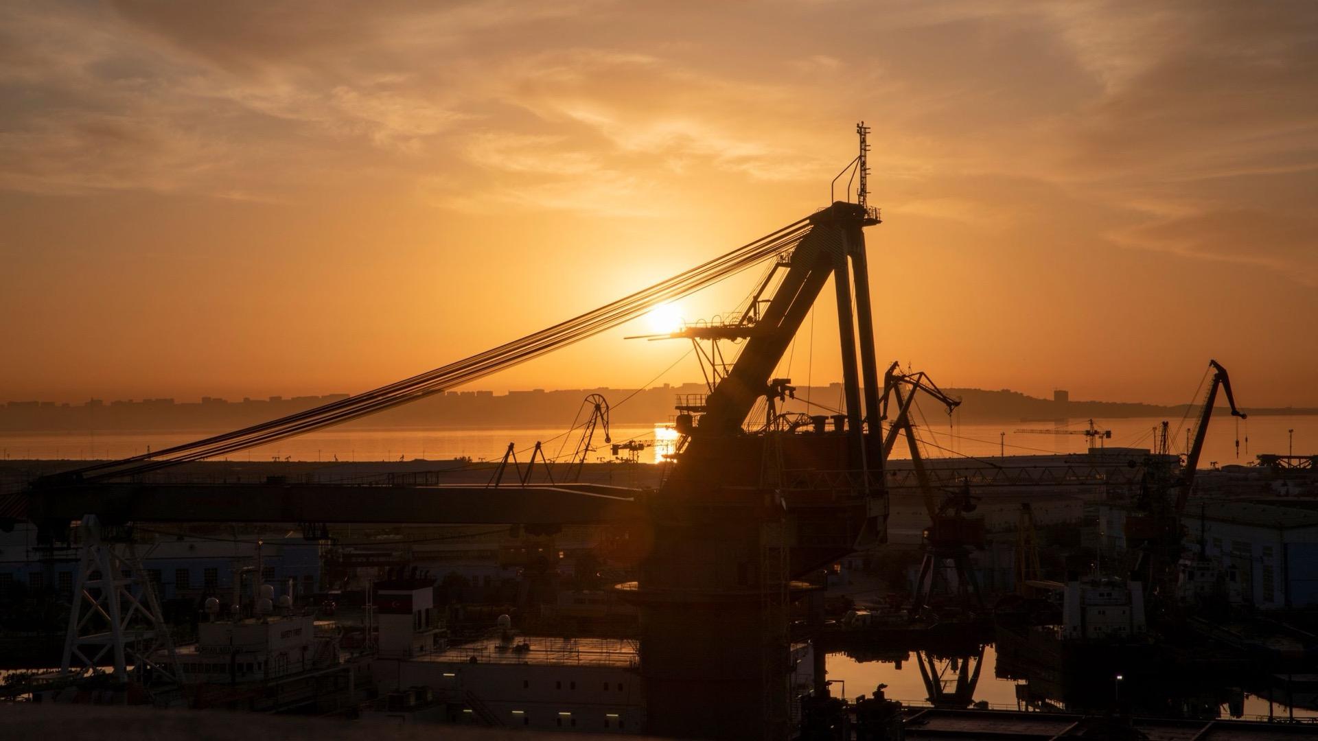 Harbour cranes in silhouette against a sunrise over the water.